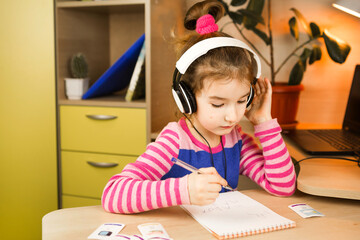 A pre-school girl with headphones is engaged at home at the table studying an educational program, writing in a notebook. Distance learning, education, language learning. Copy space