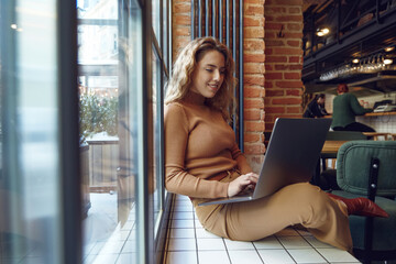 Woman sitting on windowsill of cafe with laptop on knees