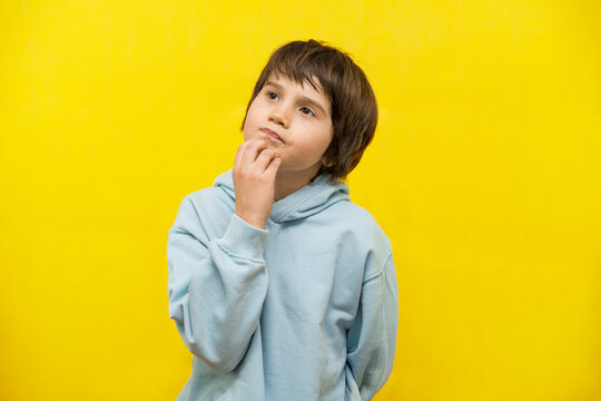 Close Up Portrait Of A School Boy Of Nine Years Old Wearing Light Blue Hoodie, Looking Up Thoughtfully And Reflectively. Child Thinking With A Hand On His Mouth. Isolated On Yellow Background.