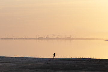 Tranquil minimalist landscape with lonely girl on calm water and industrial zone with factories and wind turbines on background. Minimal landscape photography