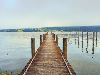Fototapeta premium A jetty of wooden planks protrudes into Lake Constance (Germany). The almost light turquoise evening sky is reflected in the water, on the opposite side you can see a strip of land.