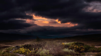 Naklejka na ściany i meble Extreme weather and lighting conditions during sunset in the Scottish Highlands.Dark and dramatic sky with black clouds with a window of orange sunlight over hills.Beautiful landscape of Scotland.