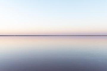 Tranquil minimalist landscape with smooth surface of the pink salt lake with calm water with horizon with clear sky in sunrise time. Simple beautiful natural calm background