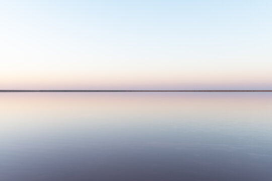 Tranquil minimalist landscape with smooth surface of the pink salt lake with calm water with horizon with clear sky in sunrise time. Simple beautiful natural calm background