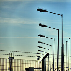 Silhouette of street lights in front of metal fence with barbed wire with light veil clouds in blue...