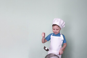 A cute little boy in a blue T-shirt, white chef's hat and apron holds a ladle and a sieve on a light green background