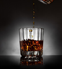 Pouring whisky into a cyrstal glass with ice cubes, circular gradation of light and fog on background with reflection on shiny black ground.