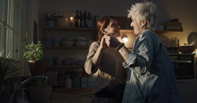 Cinematic shot of young carefree happy smiling homosexual female gay and transgender man couple in love is enjoying time together, dance together and kissing while cooking in kitchen at home.
