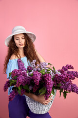 Portrait of a happy beautiful young girl with basket full of lilac on pink background.