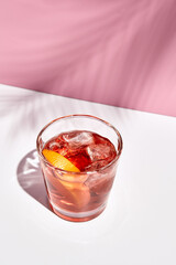 Negroni cocktail over pink background. Drink in rox glass in daylight with palm leaf hard shadow. Summer, tropical, fresh drink concept