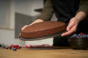 A proud cook woman shows the homemade chocolate cake she has just baked in her kitchen. She will decorate the sweet bakery with blackberries, red currants, powdered sugar and dark chocolate.