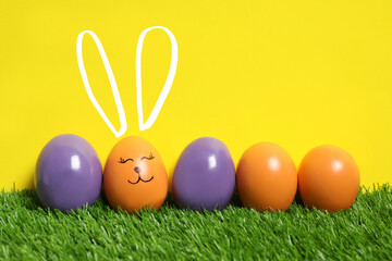 One egg with drawn face and ears as Easter bunny among others on green grass against yellow background