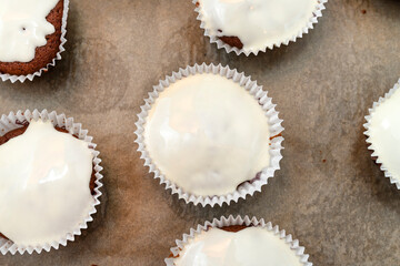 Chocolate brown muffin cake wrapped in white paper and covered with white frosting, baked in the oven, lying on baking paper, top view.