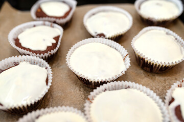 Chocolate brown muffin cake wrapped in white paper and covered with white frosting, baked in the oven, lying on baking paper.