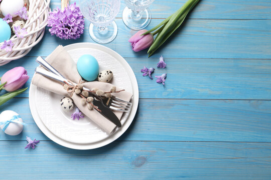 Festive Easter table setting with  painted eggs and floral decor on light blue wooden background, flat lay. Space for text