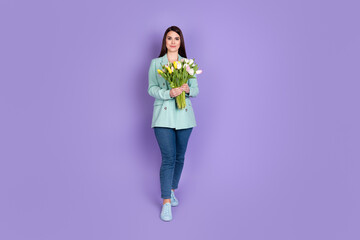 Full length photo of attractive positive woman hold flowers wear jeans teal blazer isolated on violet color background