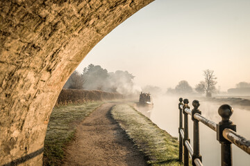 Stone bridge beautiful foggy misty canal narrow boat sunrise with smoke from chimney early morning with mist and fog rising from water as sun appears along river in countryside creating idyllic peace