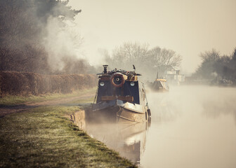 Beautiful foggy misty canal narrow boat sunrise with smoke from chimney early morning with mist and fog rising from water as sun appears along river in countryside creating idyllic peaceful atmosphere