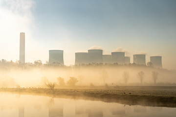 Fototapeta na wymiar Coal fired electric power station with chimneys and cooling towers surrounded by fog and mist at sunrise next to river creating beautiful orange foggy haze 