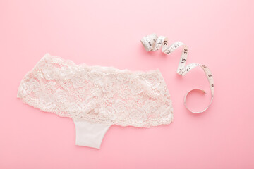 White measure tape and lace panties on light pink table background. Pastel color. Closeup. Choosing right size. Top down view.