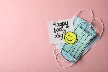 Medical masks and one paper with Happy Fools' Day note on pink background, flat lay. Space for text