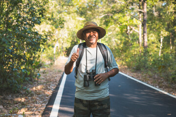 Smiling Asian elderly man with binoculars and backpack in the forest alone. Old man hiking on vacation
