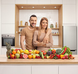 Couple posing in a kitchen behind a counter with a pile of fresh fruits and vegetables