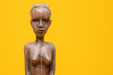 Closeup of head and torso of carved out female figurine of ebony wood without clothes on holding hands in front of her private area against a seamless yellow background.