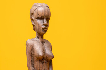 Sideview closeup of female figurine carved out of ebony wood without clothes on holding hands in front of her private area against a seamless yellow background.