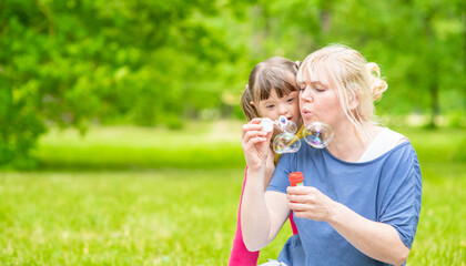 Little girl with syndrome down and her mother blow bubbles in a summer park. Empty space for text