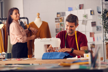 Male Fashion Designer Working On Sewing Machine As Female Colleague Works In Background