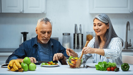 smiling asian woman mixing salad near elderly husband having breakfast in kitchen