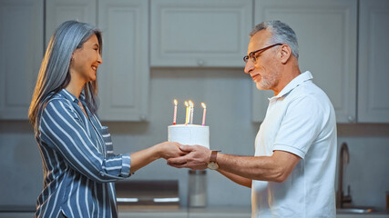 Fototapeta na wymiar side view of pleased senior man taking birthday cake from happy asian wife