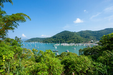 Obraz premium Marina repleta de barcos em Ubatuba, litoral de São Paulo. Pier com muitas lanchas, veleiros e embarcações ancoradas. Dia ensolarado com céu azul e poucas nuvens, sem pessoas. Árvores e montanhas.