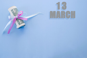 calendar date on blue background with rolled up dollar bills pinned by blue and pink ribbon with copy space. March 13 is the thirteenth day of the month