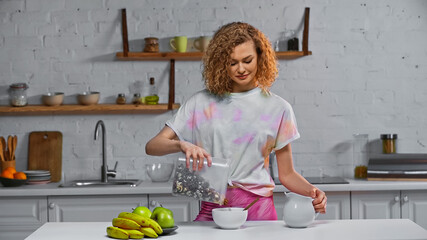 happy young adding corn flakes in bowl near fruits on kitchen table