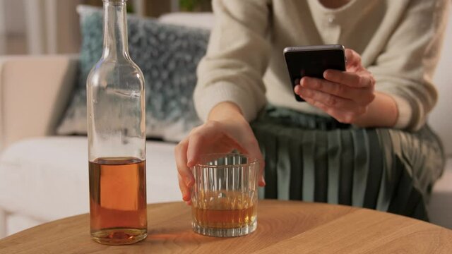alcoholism, alcohol addiction and people concept - drunk woman or female alcoholic with smartphone drinking whiskey at home
