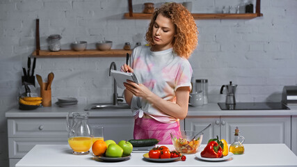curly woman writing in notebook and weighing cucumber near vegetables and orange juice on table