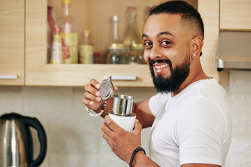Portrait of happy excited young bearded man standing in kitchen with aluminum coffee dripper