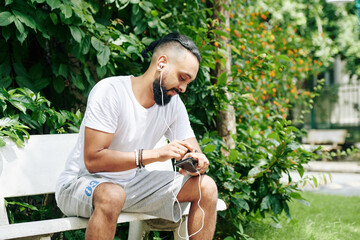 Handsome man sitting on bench in park, listening to music on smartphone and checking smartwatch