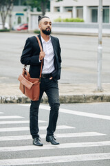 Handsome stylish bearded man with leather bag crossing road