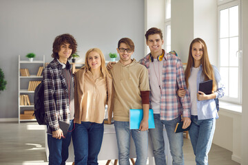 Group of happy young people in casual wear standing in modern classroom at uni. Portrait of five smart high school, college or university students smiling and looking at camera all together