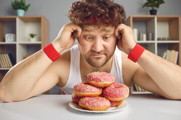 Funny athlete tempted by delicious doughnuts forgets about sports workout. Hungry man who loves sweet unhealthy junk foods sitting at table and looking at tempting fatty donuts with sprinkles on plate