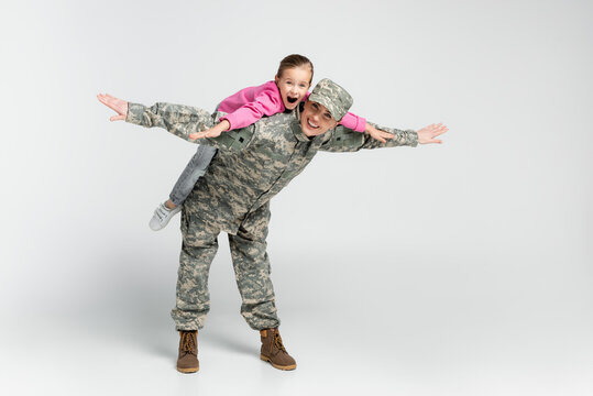 Amazed Girl Piggybacking On Mother In Camouflage On Grey Background