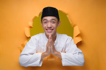 Asian muslim man smiling and shows greeting gesture poses through torn yellow paper hole, welcoming gesture to celebration of Ramadan or Eid Al-Fitr.