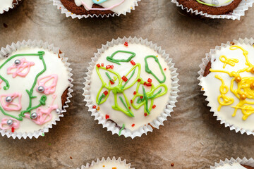 Chocolate brown muffins wrapped in white paper and covered with white frosting with colorful decorations, baked in the oven, lying on baking paper, top view.