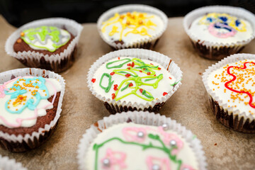 Chocolate brown muffins wrapped in white paper and covered with white frosting with colorful decorations, baked in the oven, lying on baking paper.