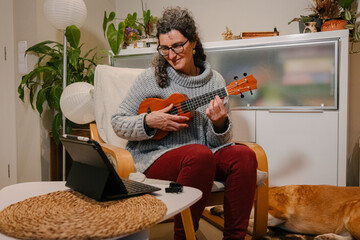Mujer caucásica de mediana edad apreniendo a tocar el ukelele en su casa con una tablet junto a su...