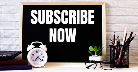 The words SUBSCRIBE NOW is written on the chalkboard next to the white alarm clock, glasses, potted plant, and pencils in a stand.