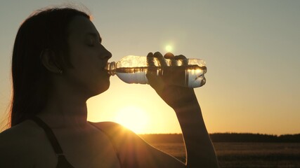 Young woman drinks water from a bottle after a workout. Jogging outdoors in rays of sunset. Outdoor sports in the park in summer, spring, girl drinks clear cool mineral water after training.
